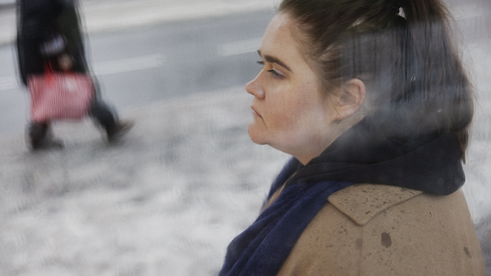 Henkilö seisoo ulkona ja katsoo vasemmalle. Taustalla ohikulkija.