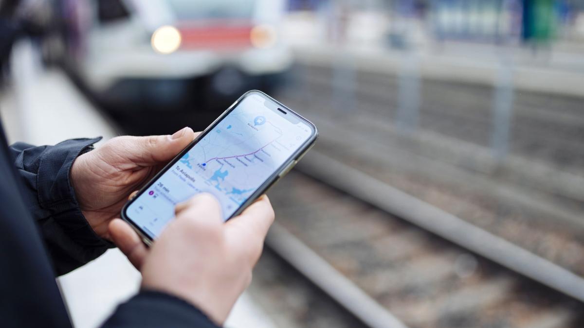 Lähikuva ihmisestä käyttämässä reittisovellusta, taustalla näkyy junaraiteet ja laiturille saapuva juna.