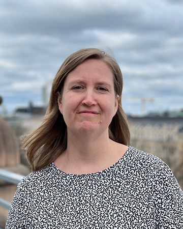 Milla Aaltonen katsoo kameraan ja hymyilee. Hänellä on yllään mustavalkoinen paita. Taustalla näkyy kaupunkimaisema.