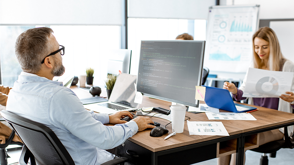 Mies selkä kameraan päin istumassa tietokoneen äärellä. Miestä vastapäätä nainen istumassa tietokoneen äärellä katsomassa papereita.